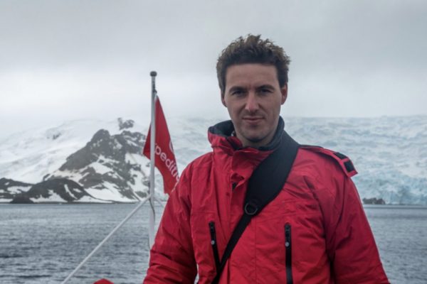 A man in a red jacket on board the deck of a ship sailing in Antarctica